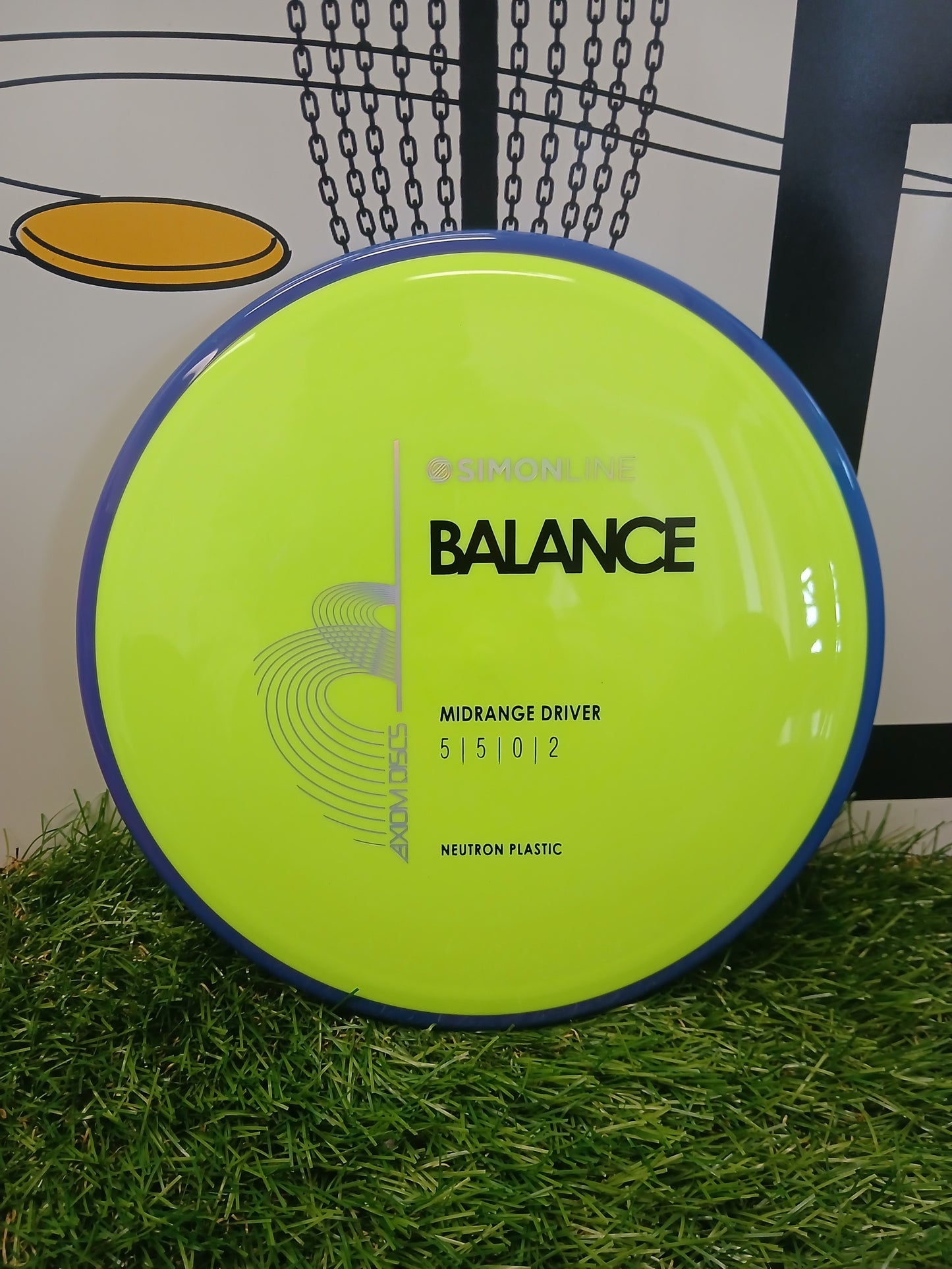 Axiom Discs SimonLine Balance midrange driver in neon yellow Neutron plastic, resting on grass near a disc golf basket.