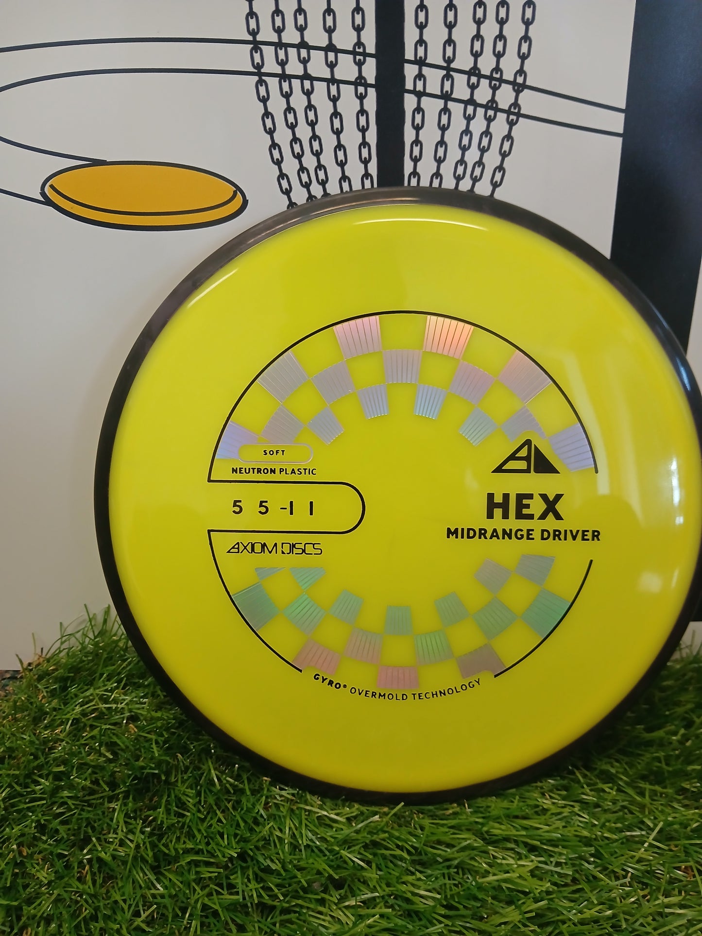 Axiom Discs HEX midrange driver in bright yellow Neutron plastic, featuring Gyro Overmold Technology, resting on artificial grass.