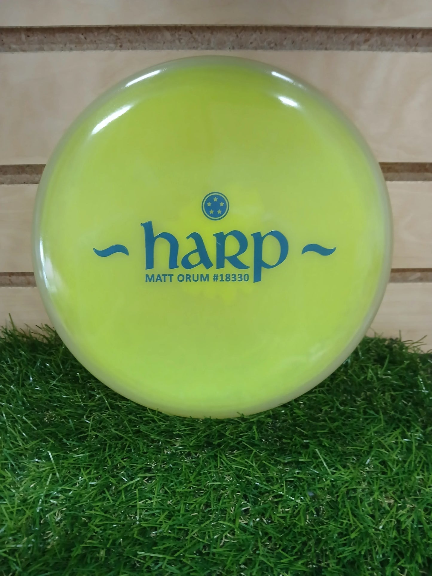 Bright yellow Harp disc golf disc by Matt Orum, resting on green grass with wooden background