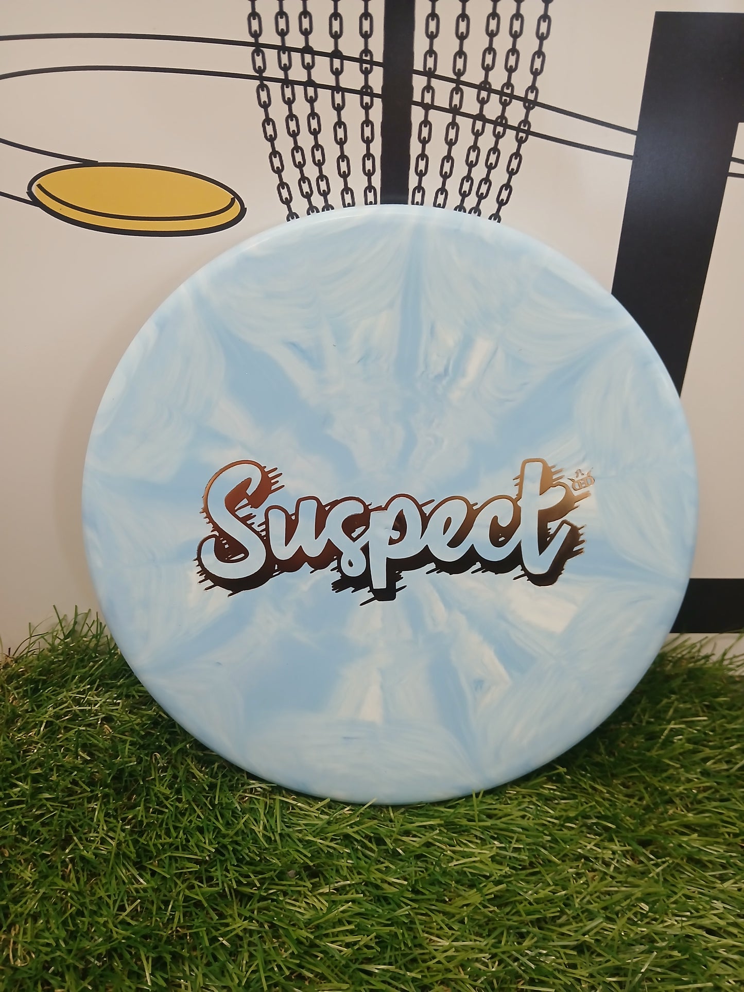 Light blue and white swirled disc golf disc with 'Suspect' logo, resting on grass near a chain basket.