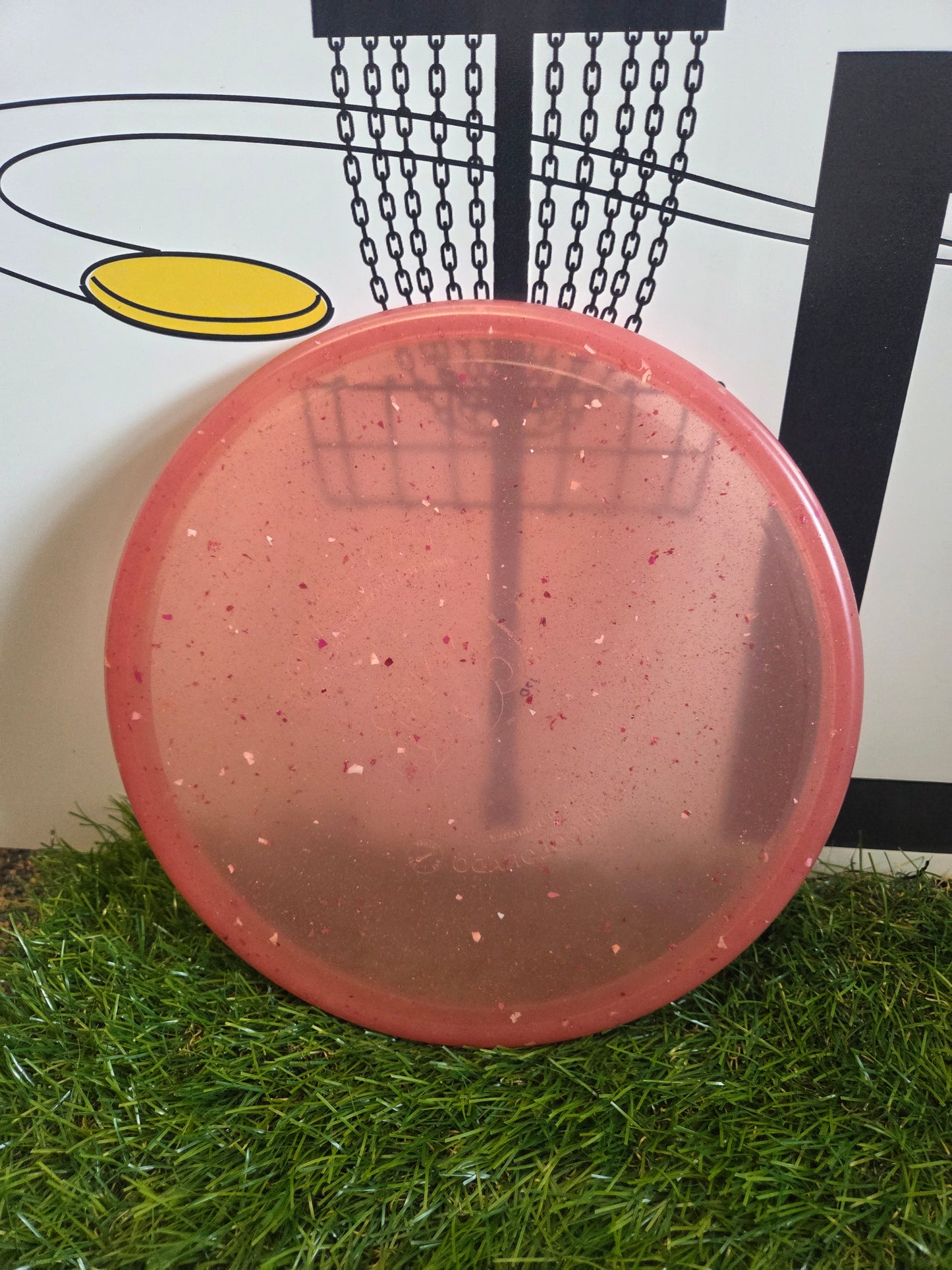 Pink speckled disc golf disc on grass with basket graphic background