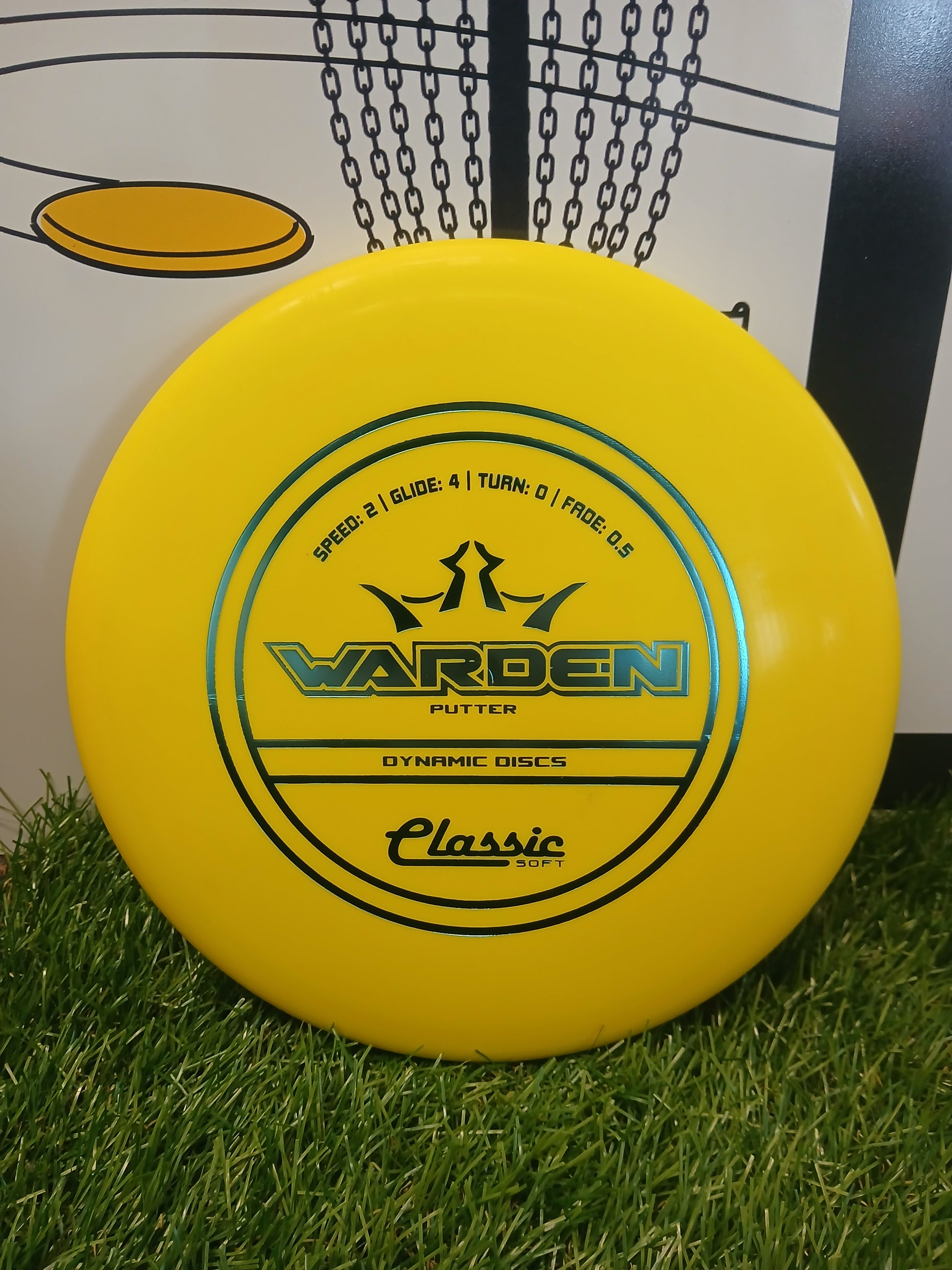 Yellow Warden putter disc by Dynamic Discs, Classic Soft, on grass near a disc golf basket.