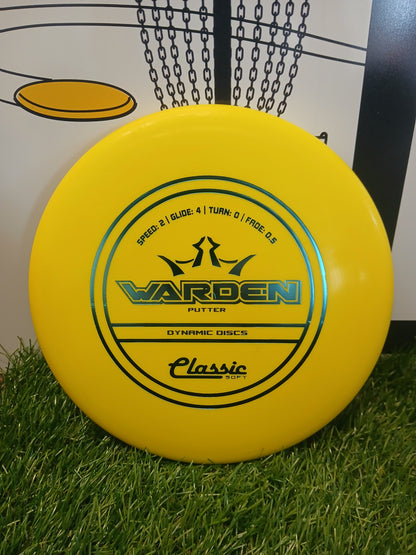 Yellow Warden putter disc by Dynamic Discs, Classic Soft, on grass near a disc golf basket.