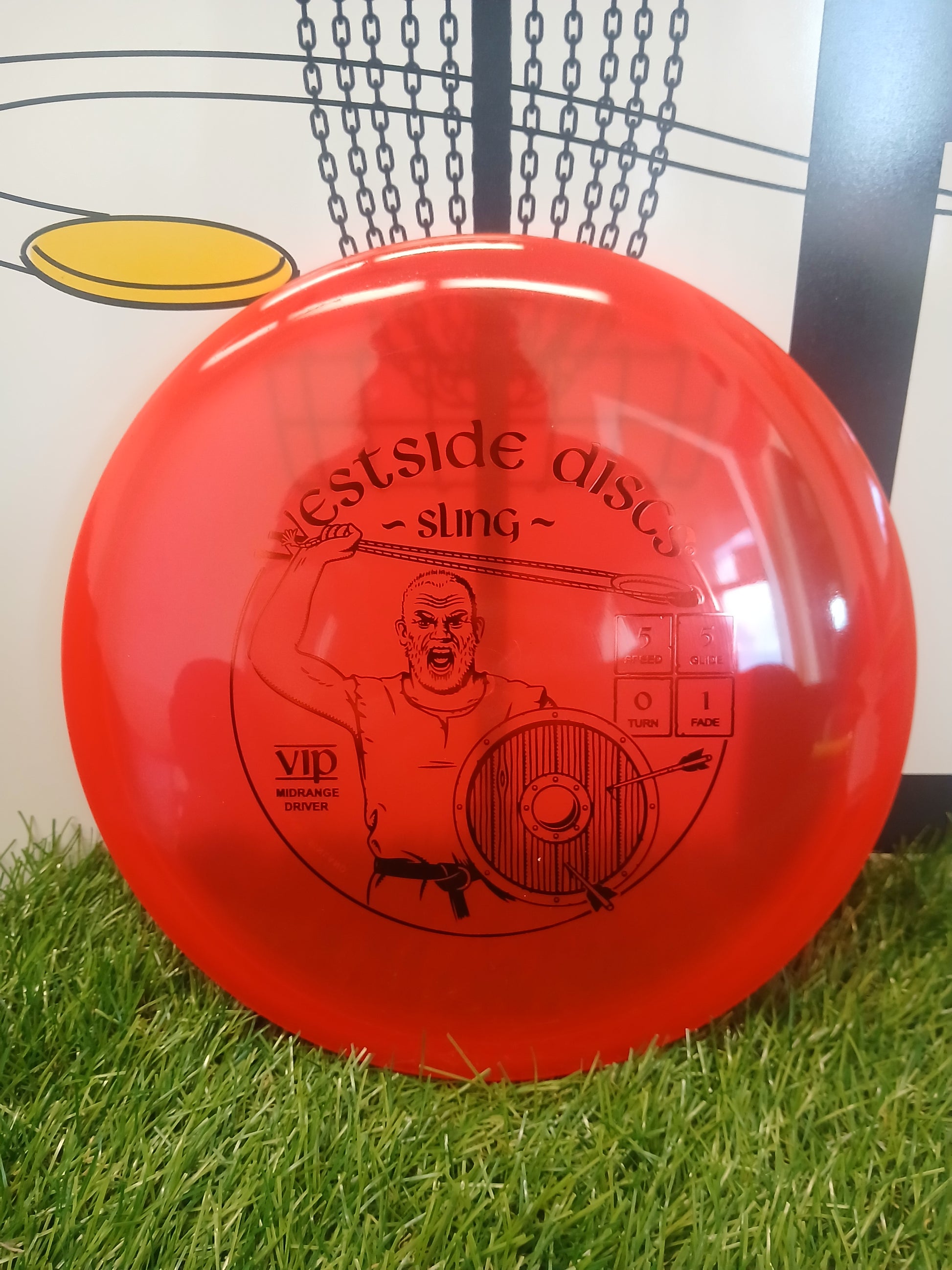 Red Westside Discs Sling VIP midrange driver disc golf disc on grass with chain-link basket background