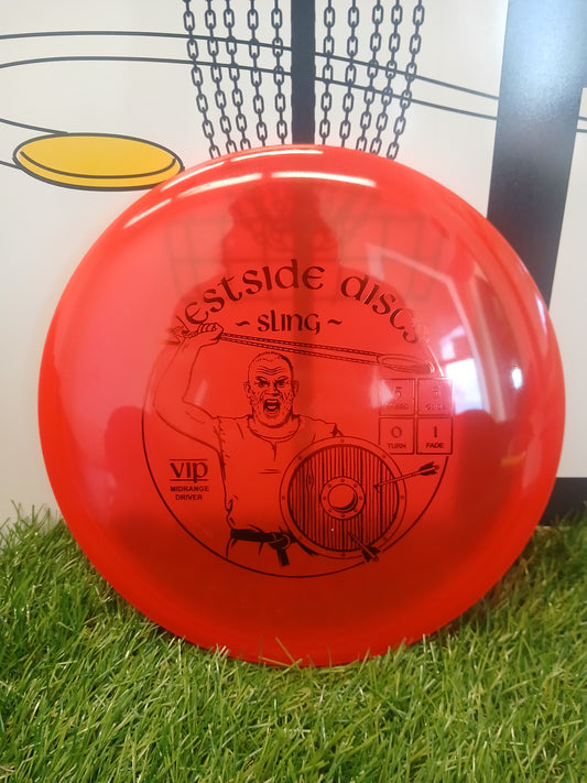 Red Westside Discs Sling VIP midrange driver disc golf disc on grass with chain-link basket background