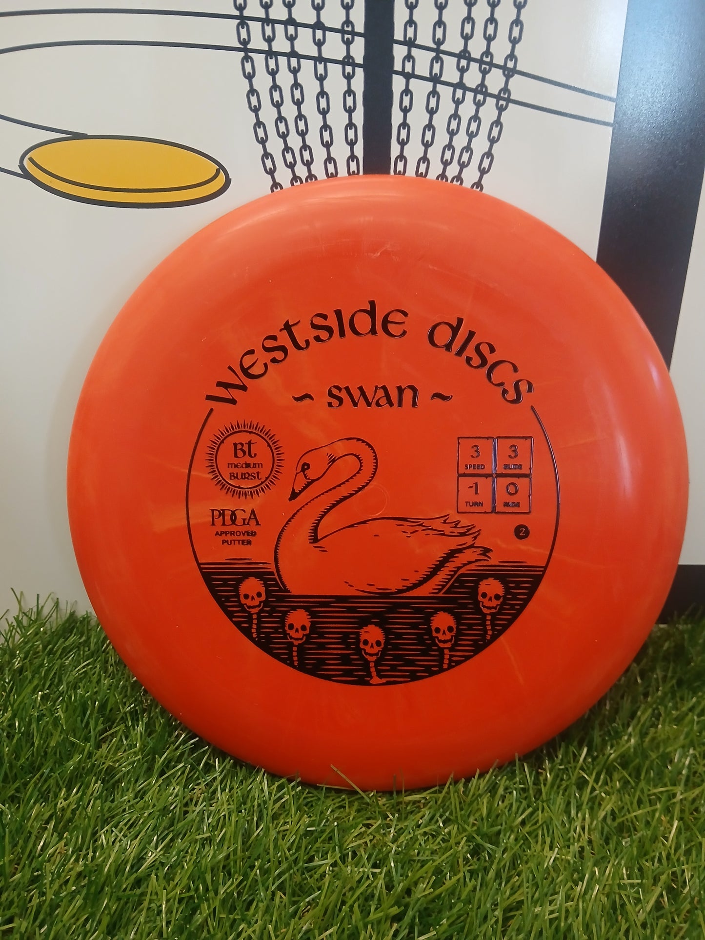 Westside Discs Swan putter disc, orange with swan graphic, PDGA approved, on grass near basket
