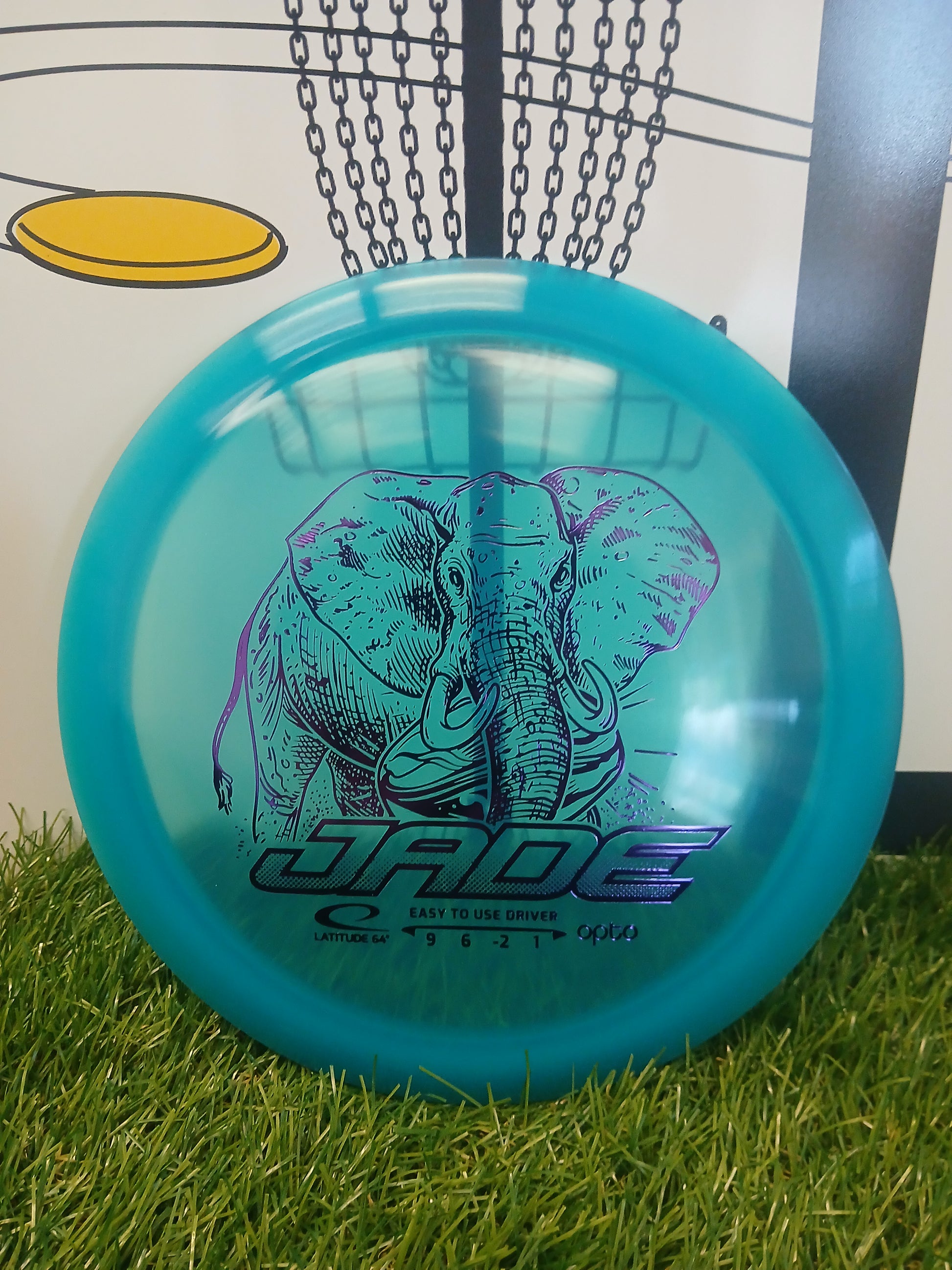 Blue Jade disc golf driver with elephant design, resting on grass near basket