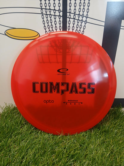 Red Latitude 64 Compass Opto midrange disc golf disc on grass with basket background