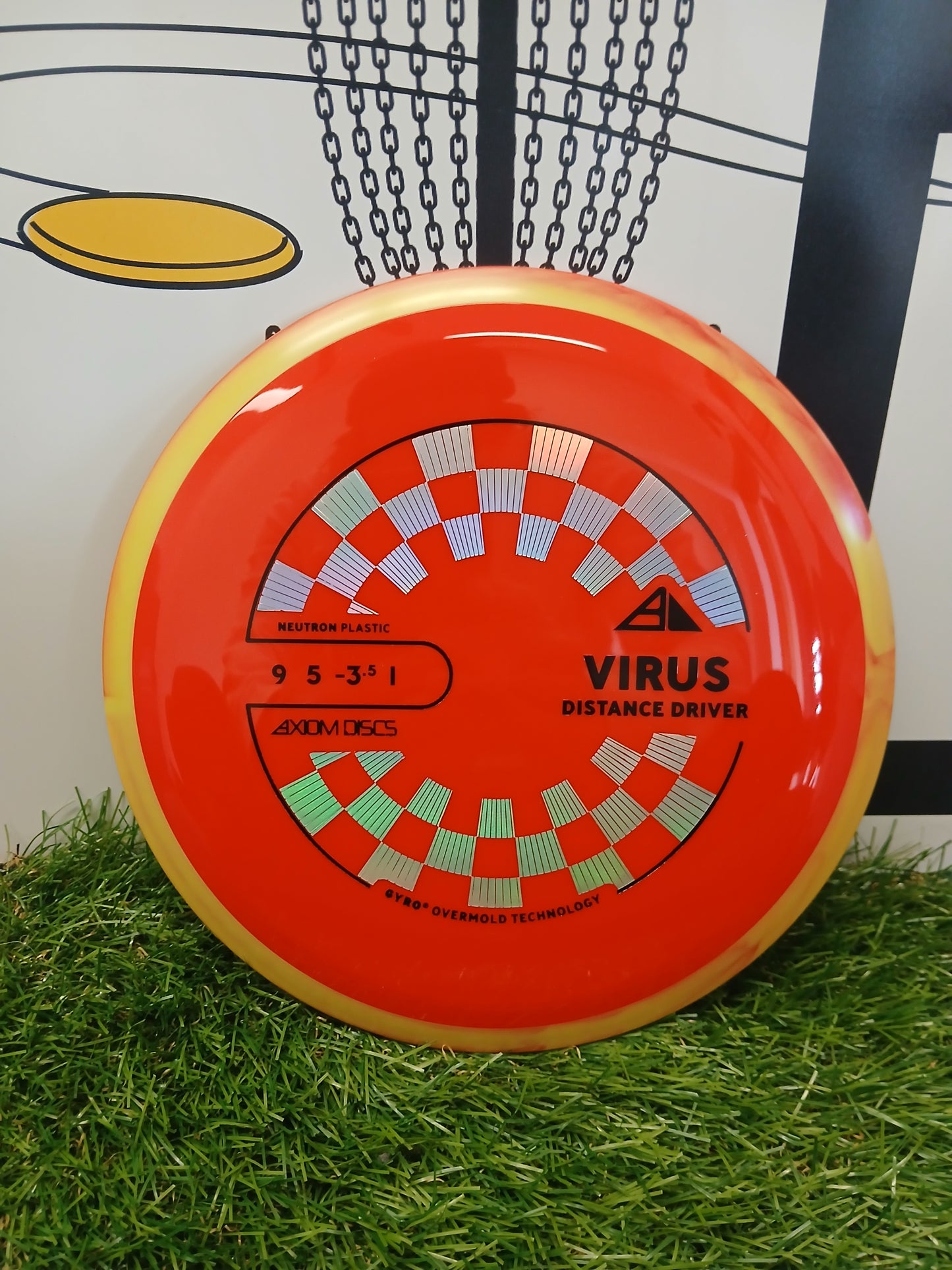 Axiom Discs Virus distance driver disc golf disc in neutron plastic, resting on grass with chain basket background.