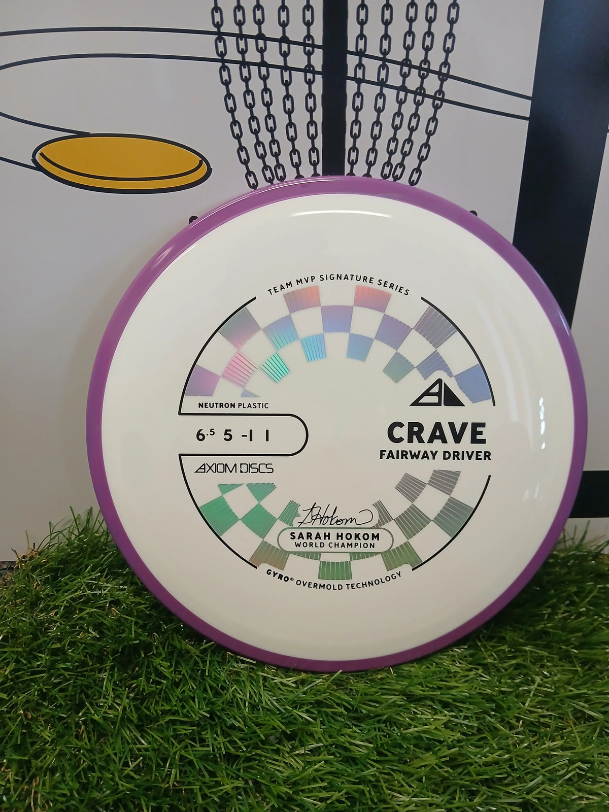 Axiom Discs Crave fairway driver in Neutron plastic, Sarah Hokom signature series, on grass with disc golf basket graphic.