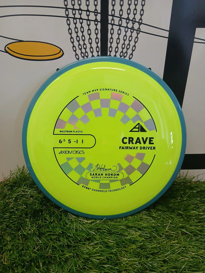 Axiom Discs Crave fairway driver in neon yellow, Team MVP Signature Series, on grass with chain-link basket background.