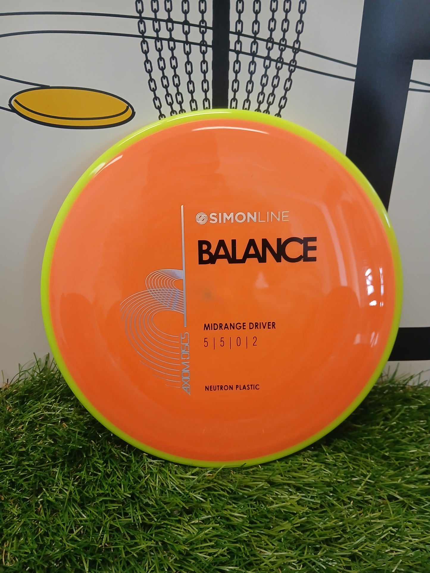 SimonLine Balance midrange driver disc golf disc in neon orange and lime green, Neutron plastic, on artificial grass near chain basket.