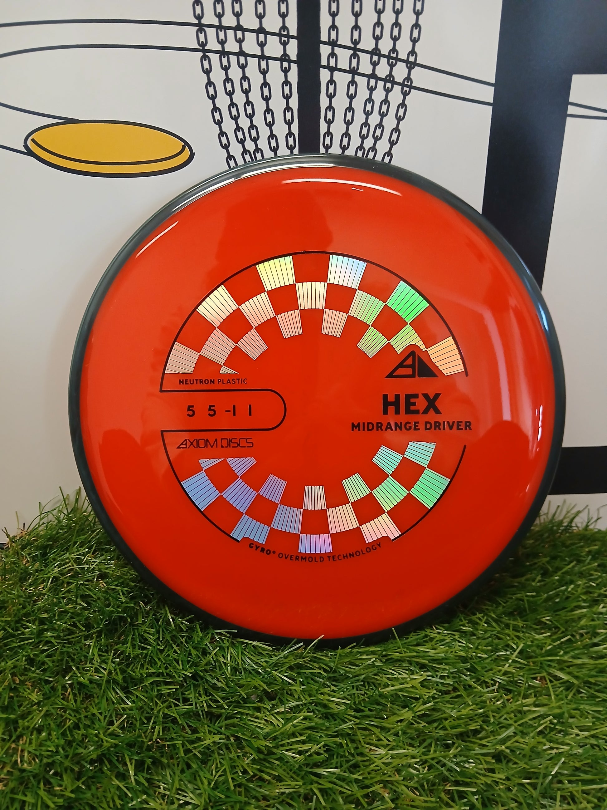 Axiom Discs HEX Midrange Driver in vibrant red with checkered design, resting on green grass near a disc golf basket.