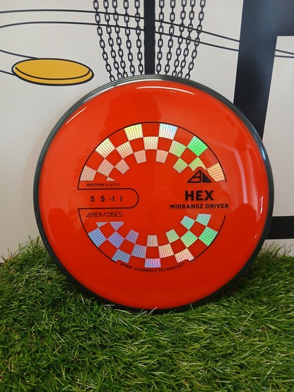 Axiom Discs HEX Midrange Driver in vibrant red with checkered design, resting on green grass near a disc golf basket.