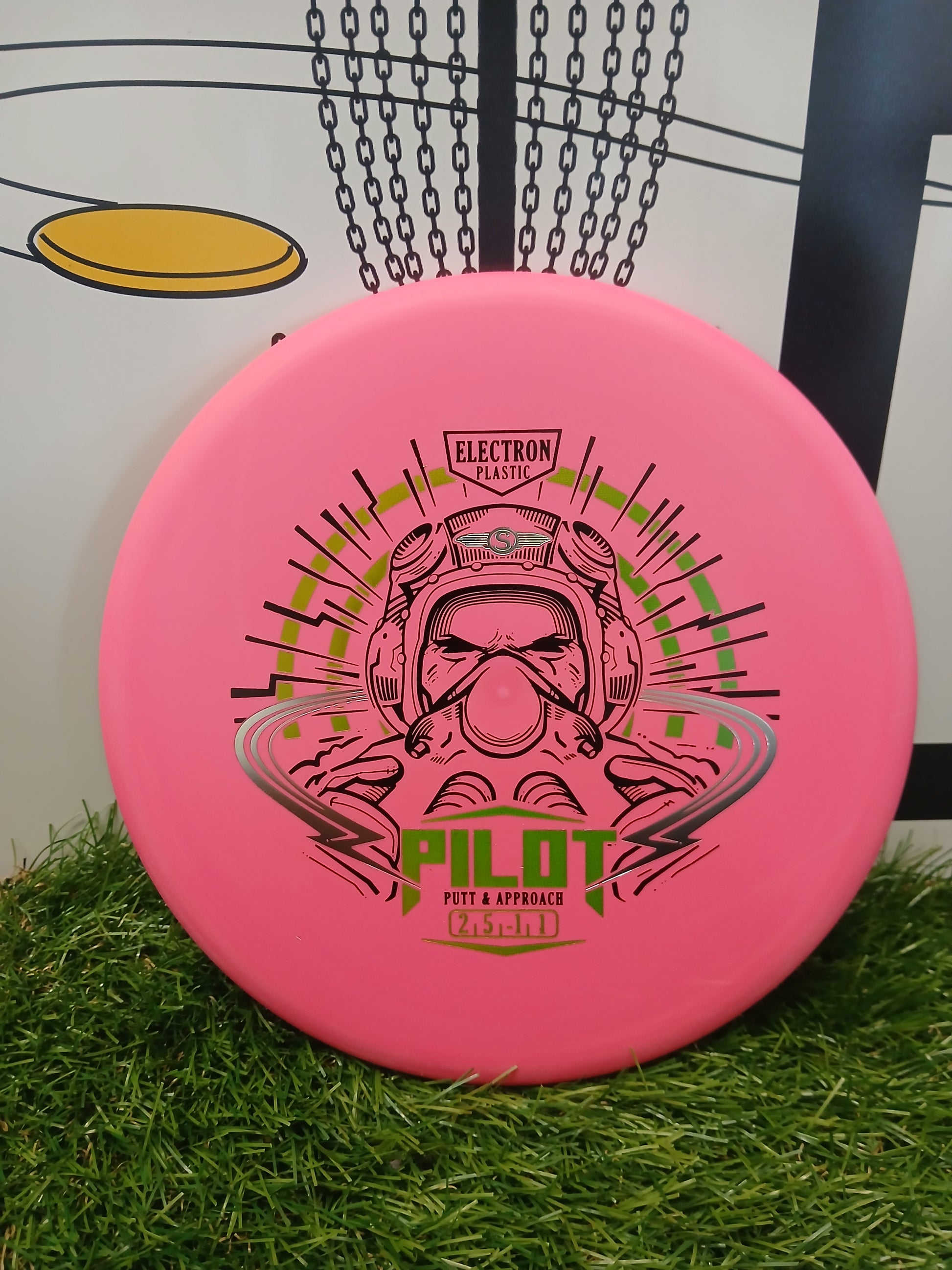 Pink Pilot putter disc golf disc with Electron Plastic, resting on grass near a basket.