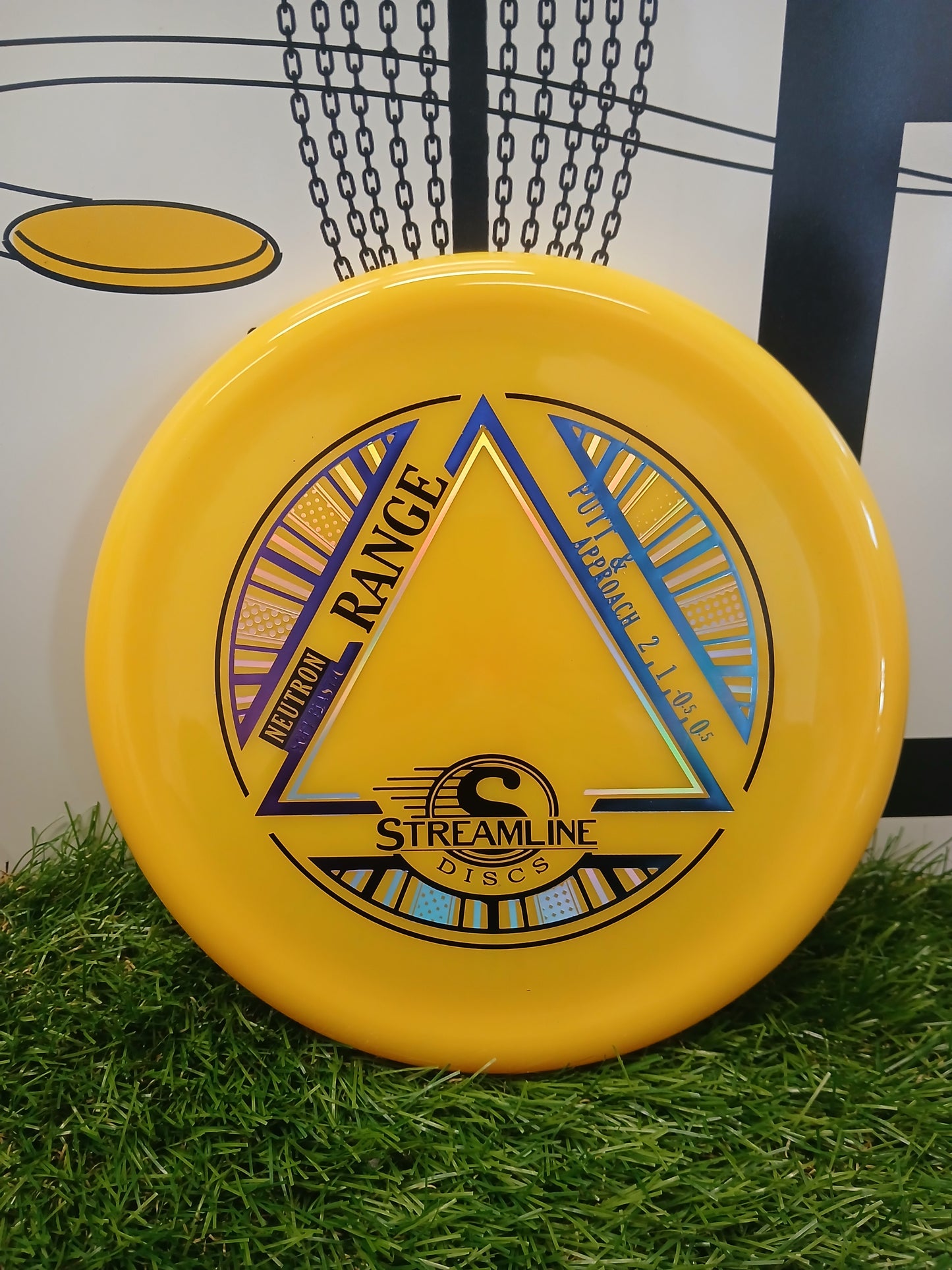 Yellow Streamline Discs Neutron Range putter for disc golf, resting on grass with basket chains in background