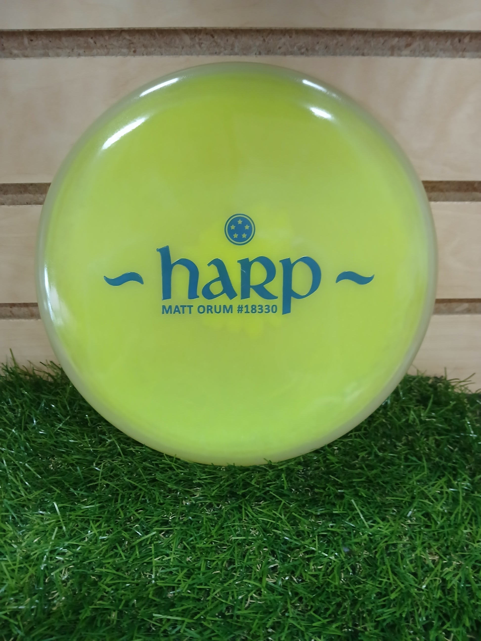 Bright yellow Harp disc golf disc by Matt Orum, resting on green grass with wooden background