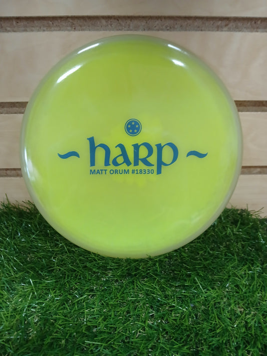 Bright yellow Harp disc golf disc by Matt Orum, resting on green grass with wooden background