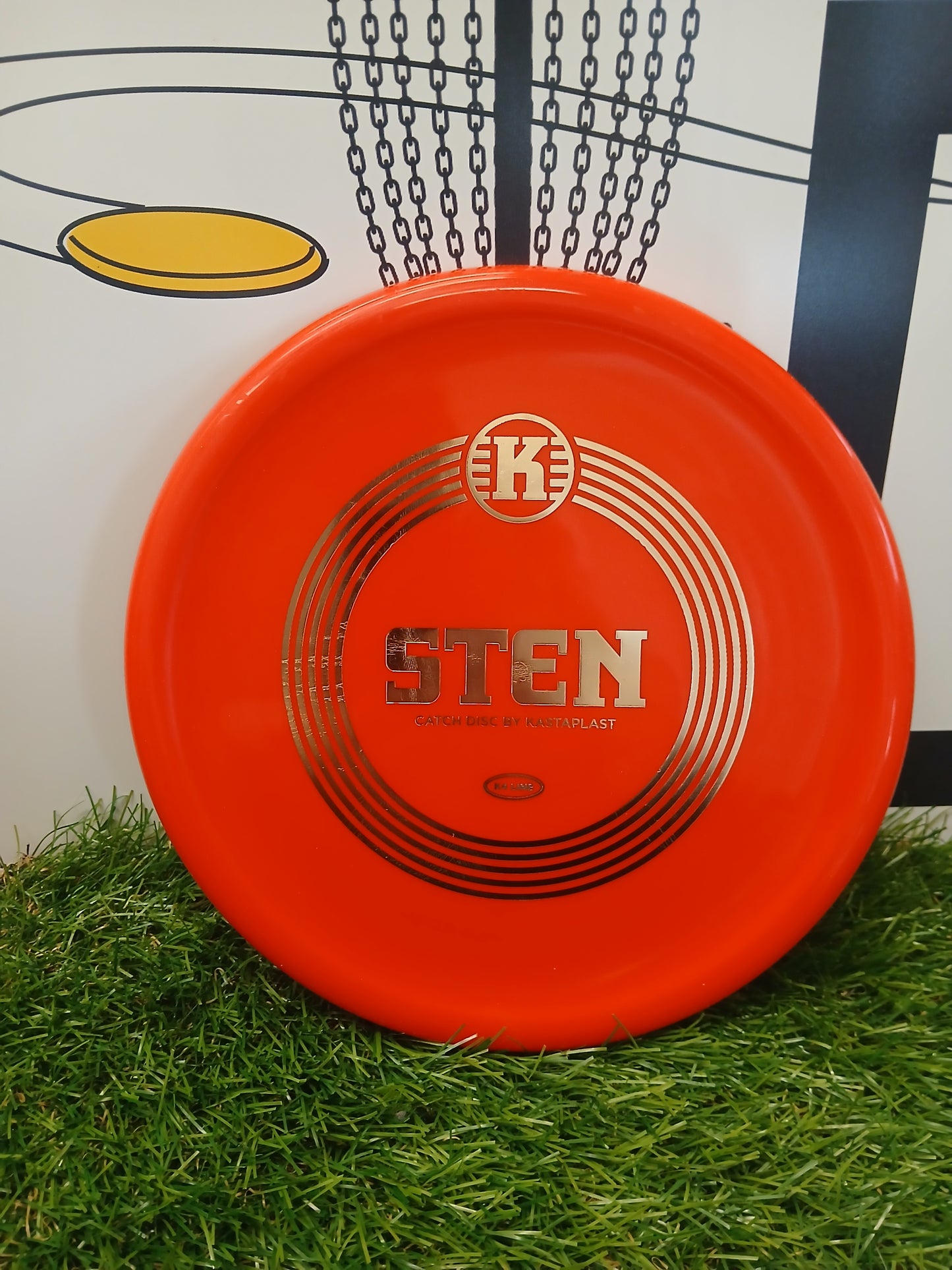 Bright orange Kastaplast Sten catch disc on artificial grass, ideal for disc golf practice and training.