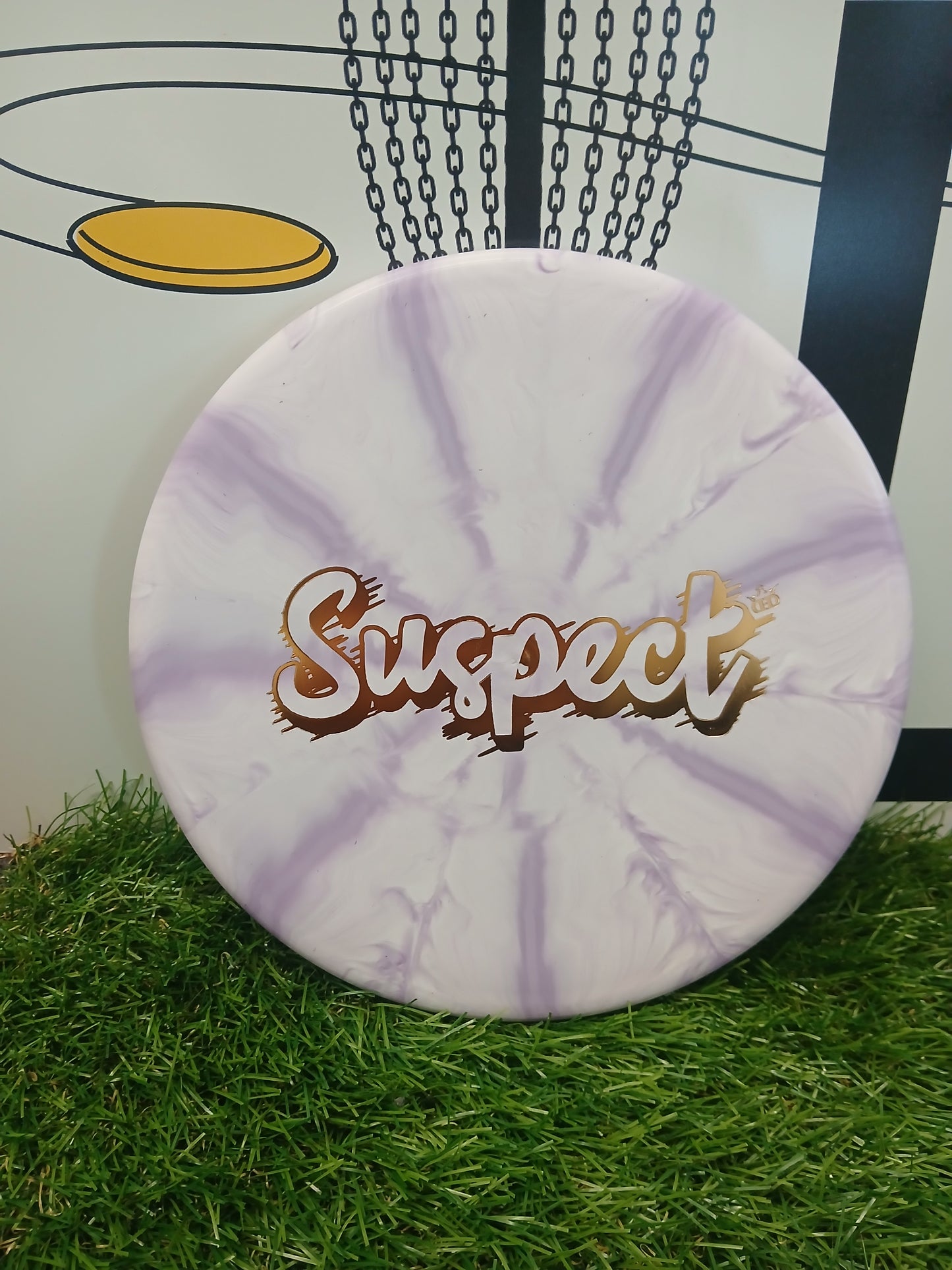 Purple and white marbled disc golf disc with 'Suspect' logo, resting on grass near a basket.