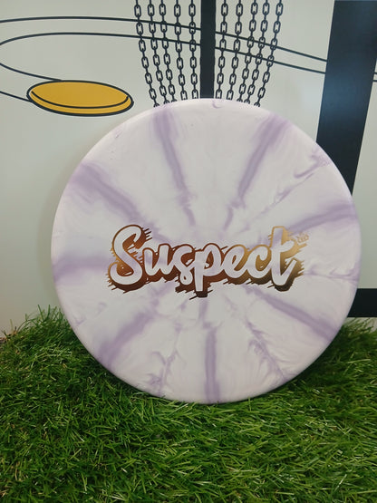 Purple and white marbled disc golf disc with 'Suspect' logo, resting on grass near a basket.