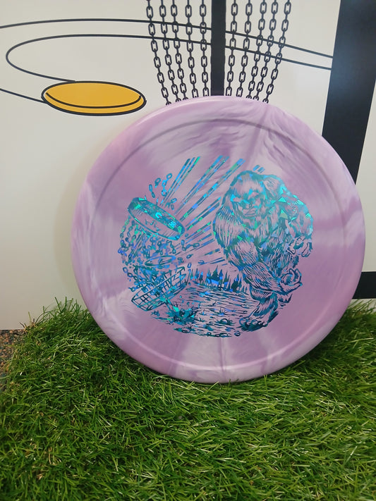 Purple disc golf disc with blue Bigfoot graphic, resting on grass near a basket.
