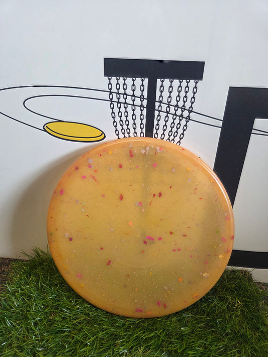 Yellow speckled disc golf disc on artificial grass with basket graphic background