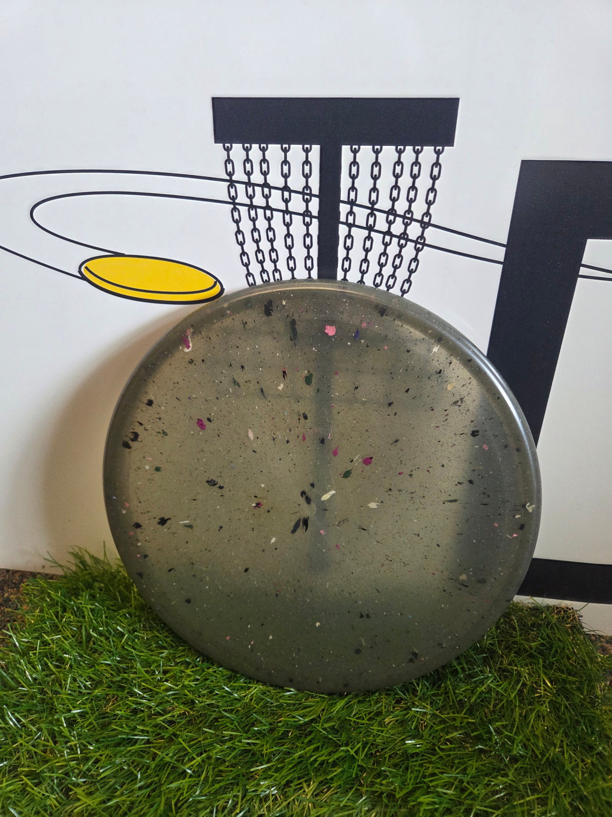 Speckled disc golf disc on grass with basket graphic background