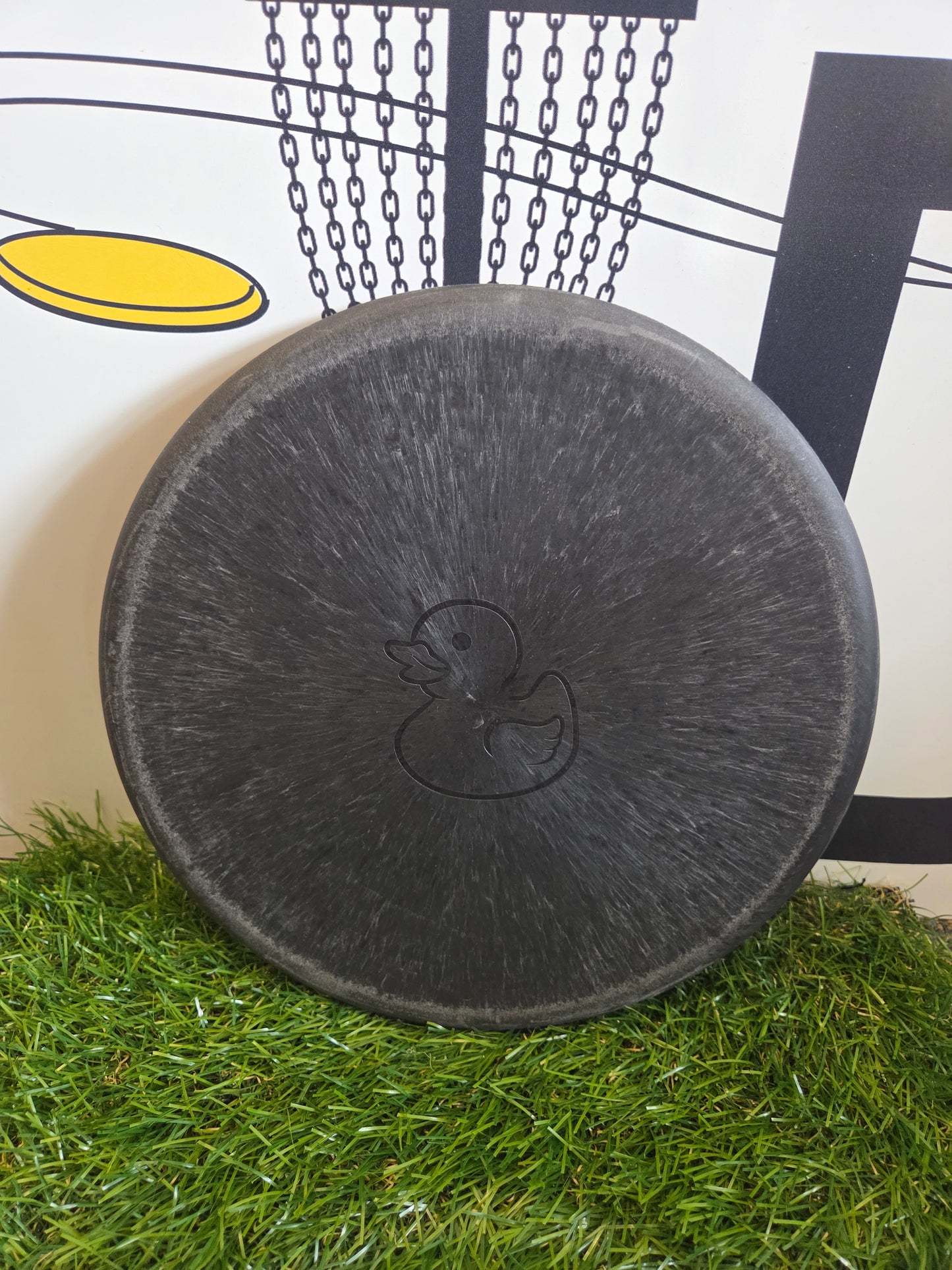 Black disc golf disc with duck logo on artificial grass, perfect for disc golf enthusiasts.