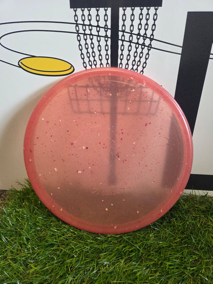 Pink speckled disc golf disc on grass with basket graphic background