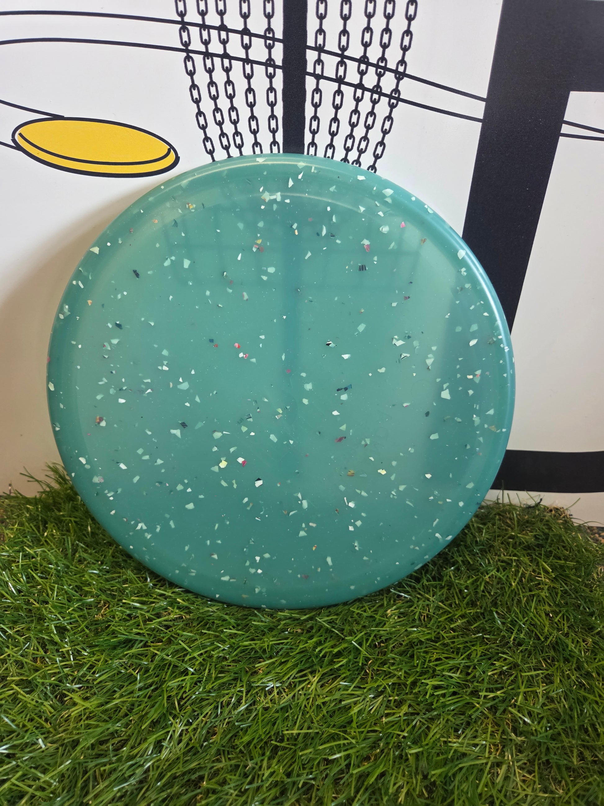Teal disc golf disc with speckled design on grass, near chain-link basket