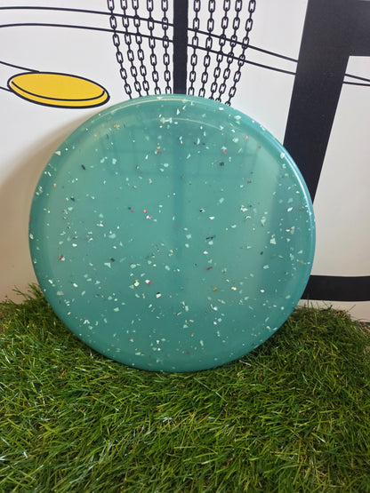 Teal disc golf disc with speckled design on grass, near chain-link basket